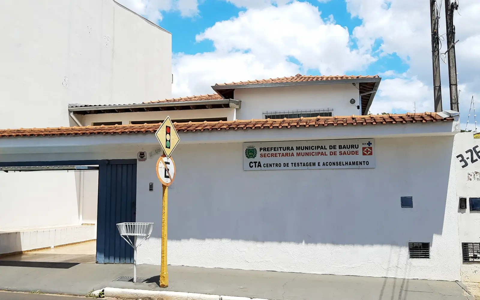 Imagem mostra fachada do Centro de Testagem e Aconselhamento de Bauru, onde funciona o Ambulatório de Assistência à Saúde de Pacientes Travestis e Transexuais. Há um muro branco, com a placa do órgão. Do lado esquerda, há uma portão de correr aberto. Atrás do muro, aparece uma parte da casa. O céu está azul, com nuvens.