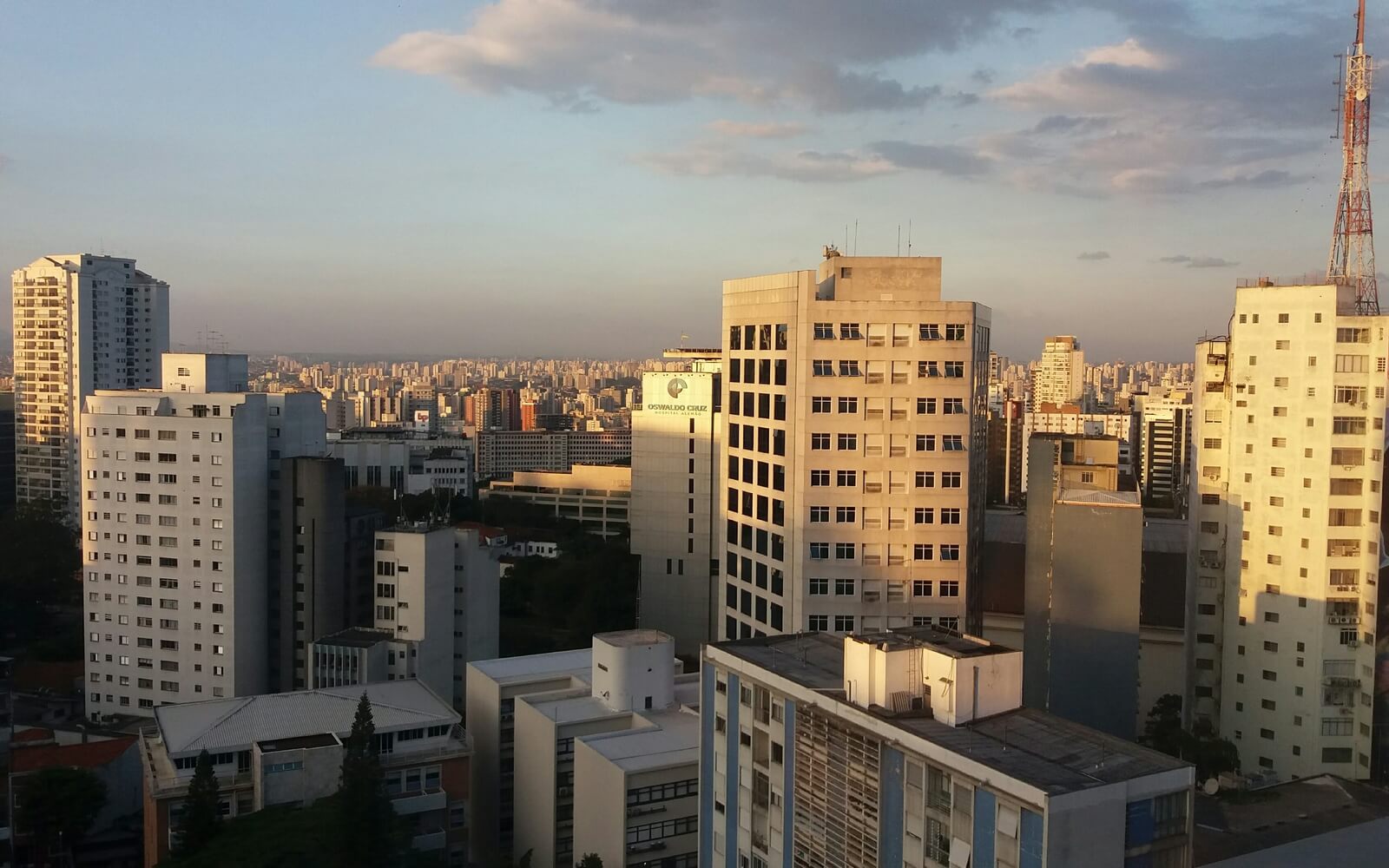 Vista de São Paulo a partir do mirante no Sesc da Av. Paulista. Há prédios em diferentes perspectivas, até o fundo da imagem. Acima, o céu tem cor degradê entre azul e cinza com algumas nuvens na direita.