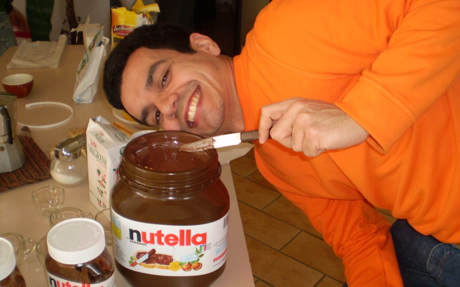 Imagem mostra Luiz Felipe curvado com o rosto próximo de um pote de Nutella grande. Luiz tem cabelo curto escuro e veste um moletom laranja.